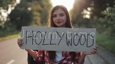 年轻漂亮的女人搭车站在路上，举着好莱坞的标志。 年轻女孩想搭车