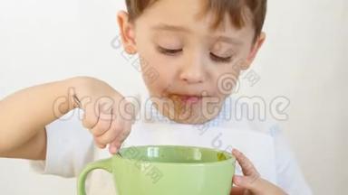 一个快乐的孩子用勺子，特写，从盘子里吃汤。 那个男孩坐在一张白色的桌子旁吃饭