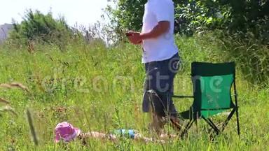 一个穿泳衣的女孩正在草坪上晒日光浴。 在她旁边的野餐椅上，一个男人坐在智能手机里玩。 他就会