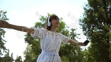 快乐的女孩戴着耳机微笑，手里拿着智能手机，在夏天的公园里跳舞和旋转。 慢动作