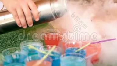 女酒保用手从热水瓶里倒出可吸烟的鸡尾酒。 多种颜色的带有管子的杯子站在绿色的背景上。