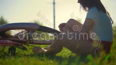 女孩坐在草<strong>地上</strong>，<strong>滚动</strong>着自行车的轮子，在后台骑着另一个女孩，阳光明媚的一天