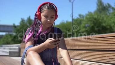女孩听音乐耳机使用智能<strong>手机</strong>坐在长凳上，心情愉快的夏<strong>季</strong>公园。 慢动作