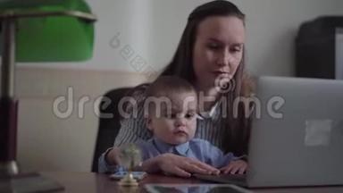 母亲和小儿子坐在室内桌子上的笔记本电脑的肖像。 忙碌的年轻母亲在家工作。 这就是