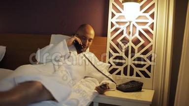 一个穿浴袍的男人躺在床上，打电话给酒店的接待处