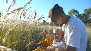 年轻的父亲手里拿着小穗，在草地上和孩子玩。 一家人坐在草地上玩得开心