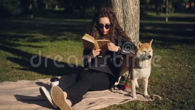 美丽的年轻女子正在看书，坐在公园树下的毯子上，微笑着，而她的家狗却在笑