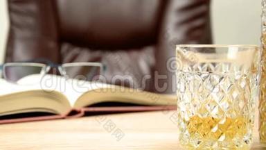 酒精饮料在水晶蜕膜。 在玻璃里，把威士忌倒在背景的木桌上，一张椅子上坐下。 开放书籍