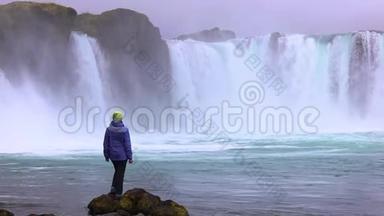一个年轻的女人欣赏一个强大的狂暴瀑布，沿着岩石边缘重<strong>重地</strong>落下。 岩石上落下一条清澈的小溪