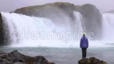 一个年轻的女人欣赏一个强大的狂暴瀑布，沿着岩石边缘重重地落下。 岩石上落下一条清澈的小溪