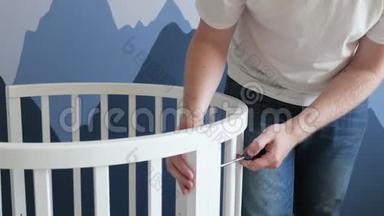 年轻人用螺丝刀为未出生的孩子收集婴儿床。 男人在墙的背景下收集婴儿床