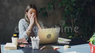 年轻的金发女人坐在办公室里用电脑工作。 她累了，所以她摸了摸她的脸和头发，摩擦