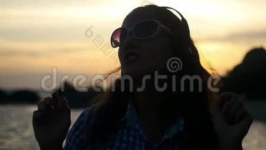 戴着耳机的年轻女子的<strong>剪影</strong>，在美丽的夕阳下，在海滩上欢快地跳舞，<strong>听</strong>音乐