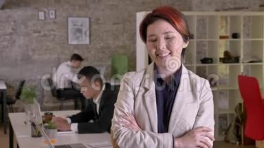 年轻的商业亚洲女人，双手交叉，在现代办公室里看着镜头，微笑着，黑人和高加索男人