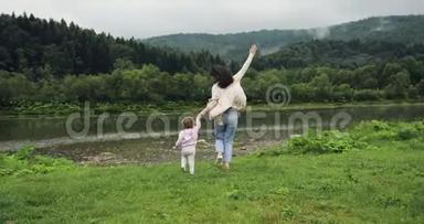 母亲带着女儿跑到河边，牵着手。 母亲的快乐时刻。 山脉背景。 风车