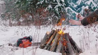 篝火晚会。 点燃了冬林中的火种.. 一个人坐在火旁，温暖他们的手。 一只链锯躺在地上