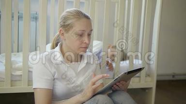 一个手里拿着电子平板电脑的女人坐在婴儿床上熟睡的小婴儿旁边