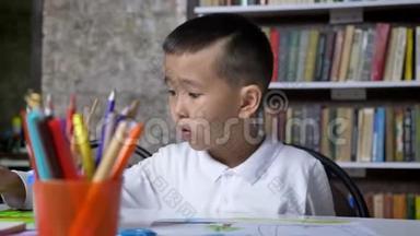 年轻的亚洲儿童集中在纸上，坐在桌子后面，男孩做作业，书架背景