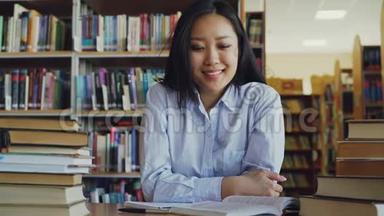 年轻漂亮的亚洲女学生坐在桌子旁，图书馆里有一堆教科书，看着摄像机