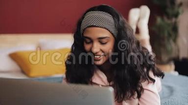 快乐的非裔美国女人躺在床上用笔记本电脑浏览网页。 穿着<strong>粉色</strong>睡衣的女孩<strong>微</strong>笑着