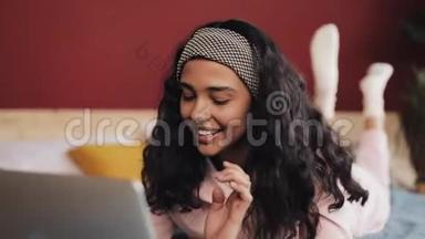 快乐的非裔美国女人躺在床上用笔记本电脑浏览网页。 穿着粉色睡衣的女孩微笑着