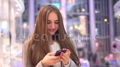 美丽的快乐女孩，长发在智能手机上写着一条信息，站在<strong>商场</strong>里。 4k