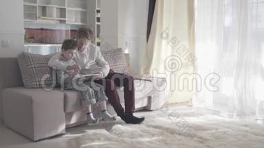 爸爸和儿子用<strong>数码</strong>平板电脑坐在大客厅的沙发上。 幸福的<strong>家</strong>庭一起度过时光.. 爸爸