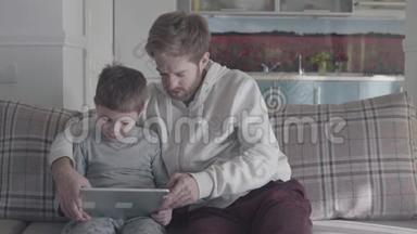父子俩坐在大客厅的沙发上玩平板电脑。 爸爸教他的孩子。 快乐的家庭开支