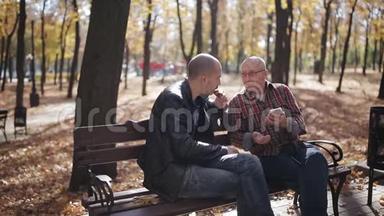 一位年迈的父亲和他成年的儿子坐在公园的长凳上喝咖啡和说话。 年长父亲的成年儿子