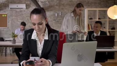 一位年轻的女商人正在办公室的智能手机上阅读和输入信息，面带微笑，她的同事们正在与
