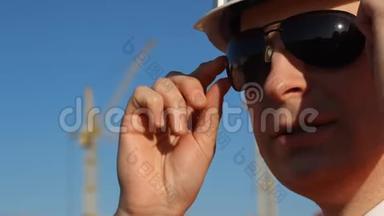 一个戴着黑色眼镜、戴着白色头盔、嚼口香糖、在建筑背景上固定眼镜的人，起重机