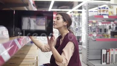 20`的漂亮女孩在电器商店里寻找微波炉，打开黑色微波炉，看看是什么