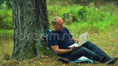 戴眼镜的人坐在公园的一棵树下，用笔记本电脑工作。