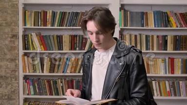 穿黑色皮夹克的年轻男学生站在图书馆看书，书架背景