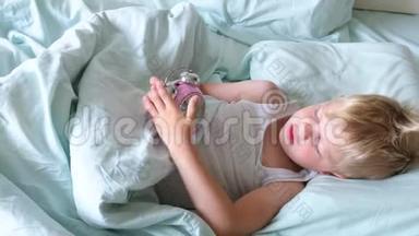 一个金发小男孩躺在床上微笑，一大早就玩闹钟。 那男孩听着钟响