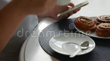 女人坐在桌子旁，喝着咖啡，夹着肉桂面包，端着咖啡，看着智能手机