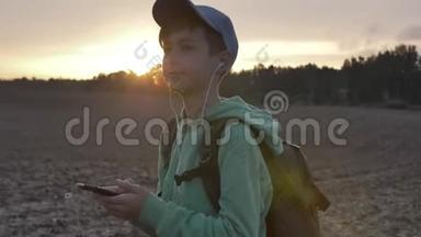 带背包的男孩在日落时走在户外，使用电话，听音乐，旅行的概念