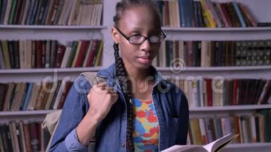年轻美丽的非洲裔美国妇女戴着眼镜，在图书馆看书，看着相机，开心地笑着