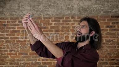 年轻漂亮的黑发男子带着胡子，正在智能<strong>手机</strong>上<strong>制作</strong>视频，四处闲逛，砖背景，交流
