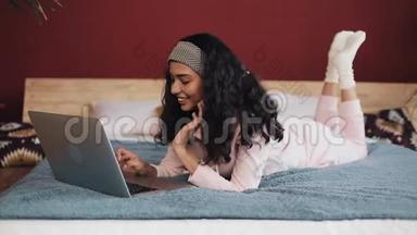 快乐的非裔美国女人躺在床上用笔记本电脑浏览网页。 穿着粉色睡衣的女孩微笑着