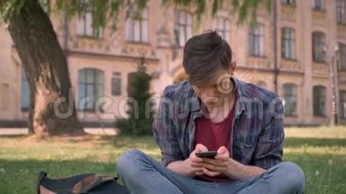 年轻的帅哥坐在公园的草地上，敲着智能手机，放松概念，交流概念，继续前进