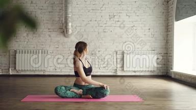 年轻的女士穿着时髦的运动服，正在做身体扭曲，坐在莲花的位置，在瑜伽工作室里的<strong>垫子</strong>上。 放松一下