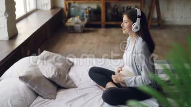微笑的怀孕女孩正在听耳机音乐，抚摸她的腹部，放松地坐在<strong>双人床</strong>上
