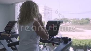 老年妇女在健身房的跑步机上订婚。 女人在电话里说话做跑步机