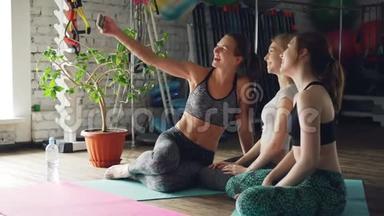 瑜伽的学生们正在用智能手机自拍，同时坐在瑜伽工作室里明亮的垫子上。 女人摆姿势