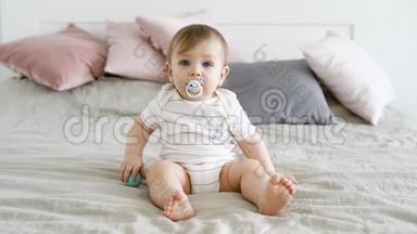 一个可爱有趣的孩子坐在床上，嘴里放着奶嘴，手里拿着球，男孩看着前方