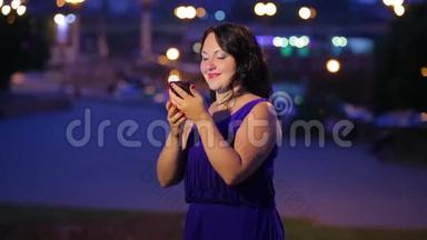 晚上，一个穿着蓝色裙子的年轻女人在街上看着智能手机上的留言，微笑着，梦幻般的样子