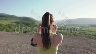 后景。 运动员女孩在日落时分在山上慢跑，从摄像机里跑出来，用耳机听音乐