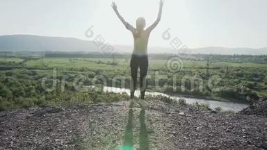 年轻的运动女孩跳起<strong>来</strong>，在日落时举起她的手在山上的岩石边<strong>缘</strong>。 快乐女孩