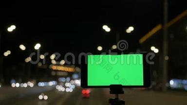 带有绿色屏幕的智能手机安装在三脚架上。 在汽车红绿灯的背景下。 晚上